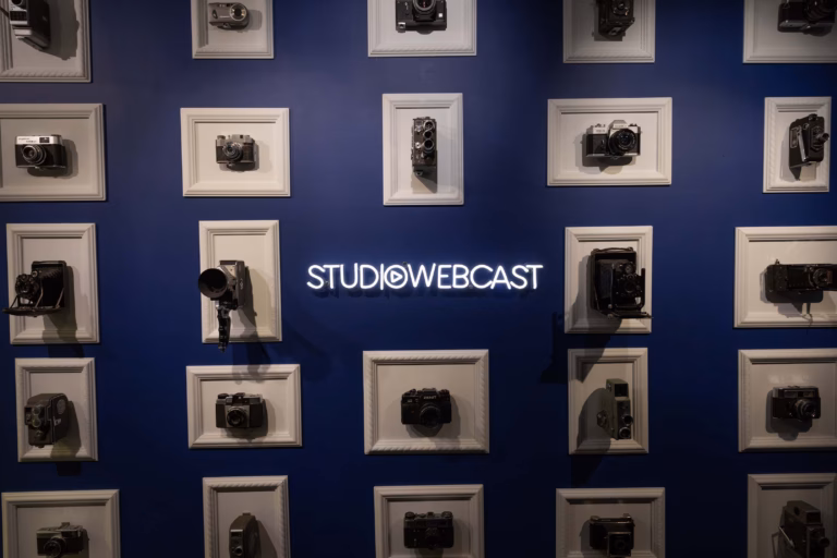 Mur décoratif de l'entrée du Studio Webcast avec une collection d'appareils photo vintage encadrés sur fond bleu nuit et le logo Studio Webcast en néon blanc