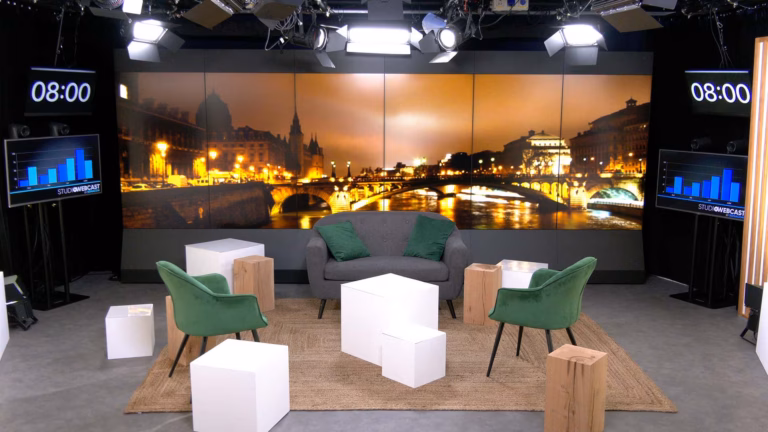 Plateau TV du Studio Webcast en configuration talk show avec fauteuils velours vert, canapé gris, poufs et cubes blancs sur fond Paris by night
