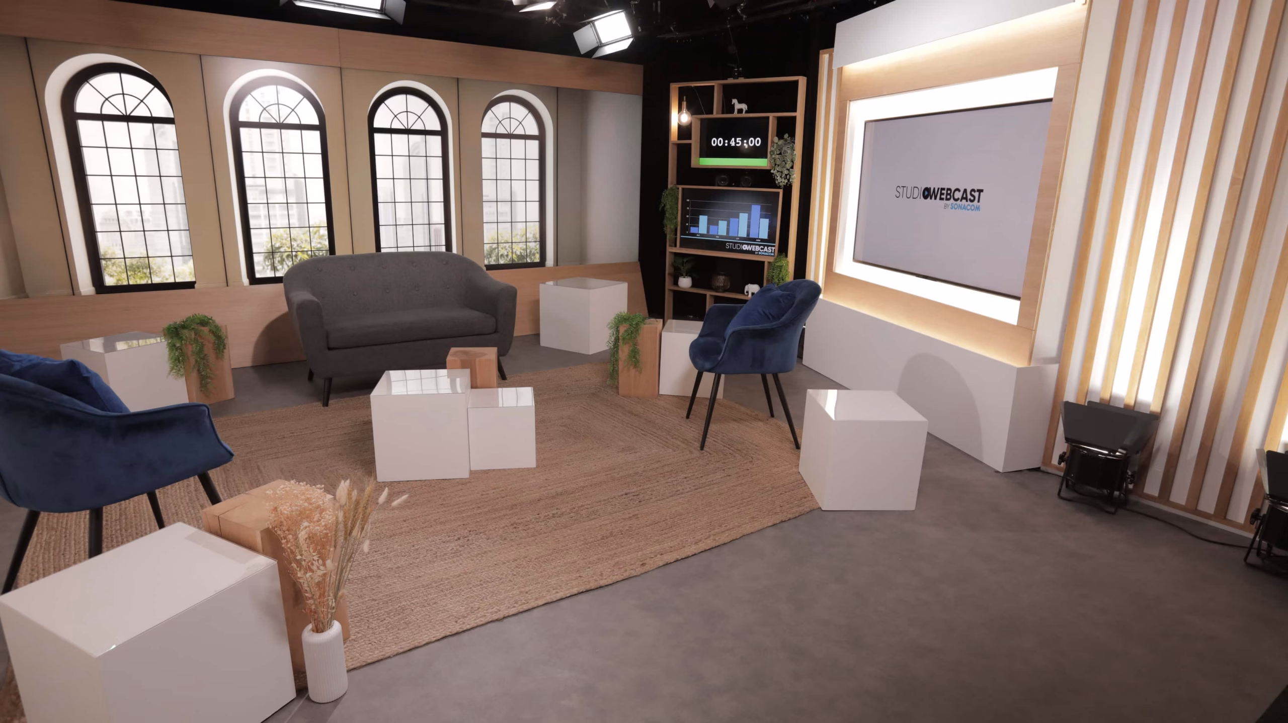Plateau TV du Studio Webcast en configuration talk show avec fond LED fenêtres, canapé gris, fauteuils bleus et cubes blancs