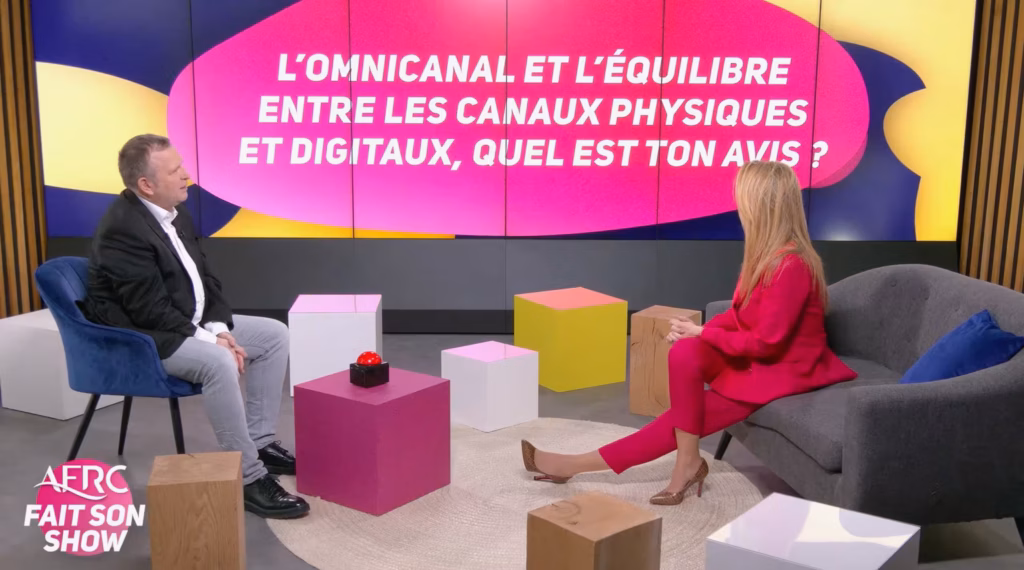 Deux intervenants en pleine discussion sur notre plateau TV et webcast à Paris. Le décor moderne, composé de fauteuils et de cubes colorés, intègre un grand écran en arrière-plan affichant une question sur l'omnicanal pour l'émission 'AFRC fait son show'.