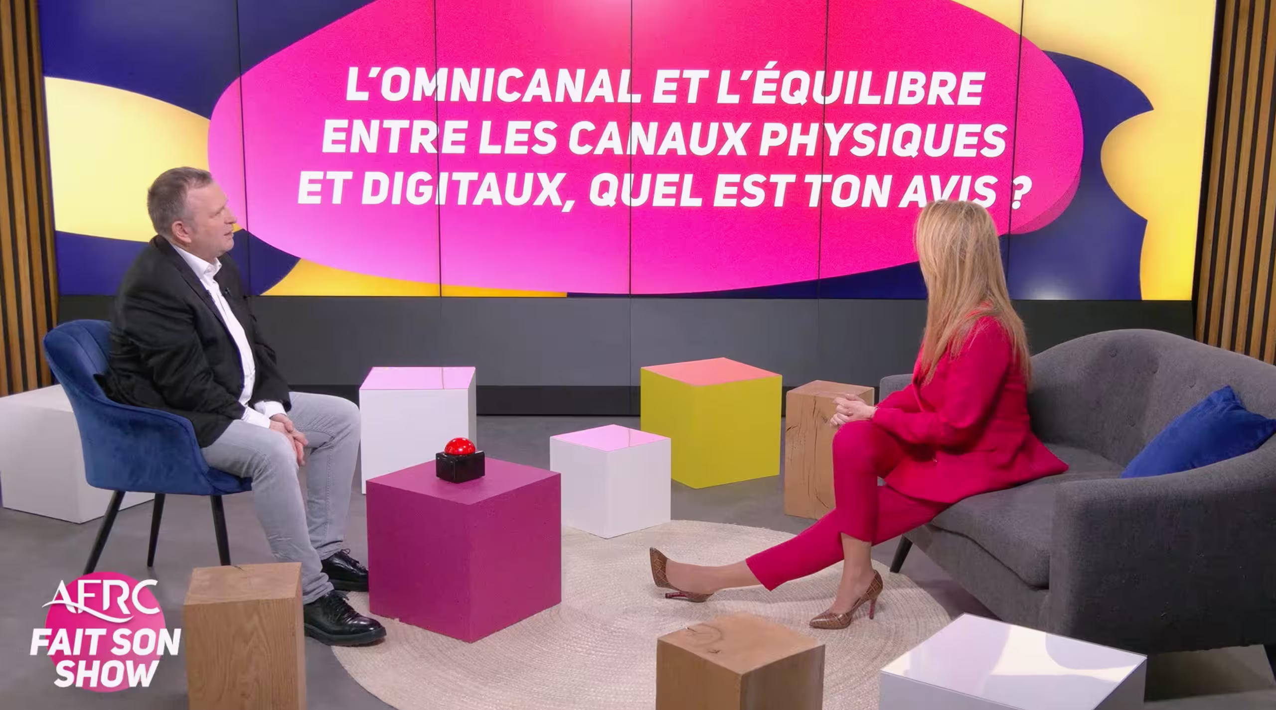 Deux intervenants en pleine discussion sur notre plateau TV et webcast à Paris. Le décor moderne, composé de fauteuils et de cubes colorés, intègre un grand écran en arrière-plan affichant une question sur l'omnicanal pour l'émission 'AFRC fait son show'.